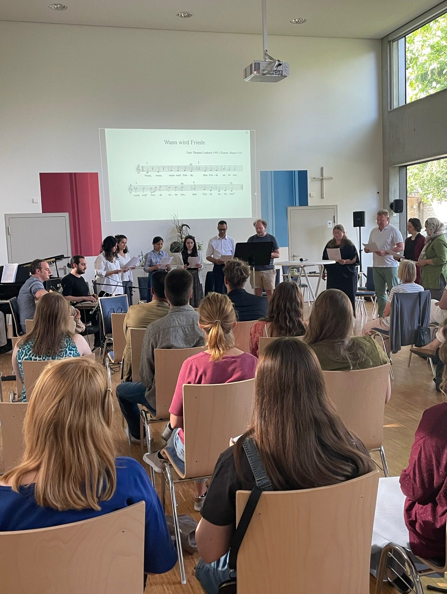 Zahlreiche Personen sitzen in einem Raum. Eine Gruppe von Personen musiziert im Vordergrund.