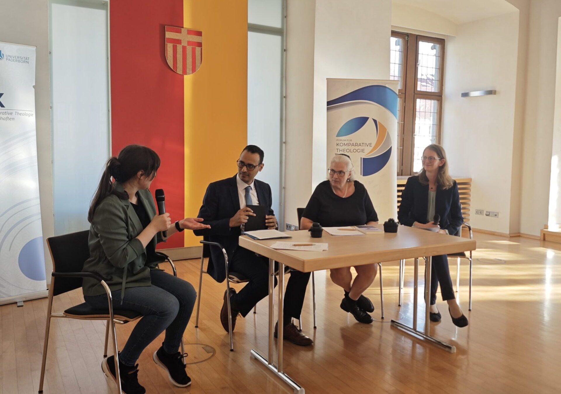 Drei Frauen und ein Mann sitzen um einen Tisch herum. Eine Frau hat ein Mikrofon in der Hand.