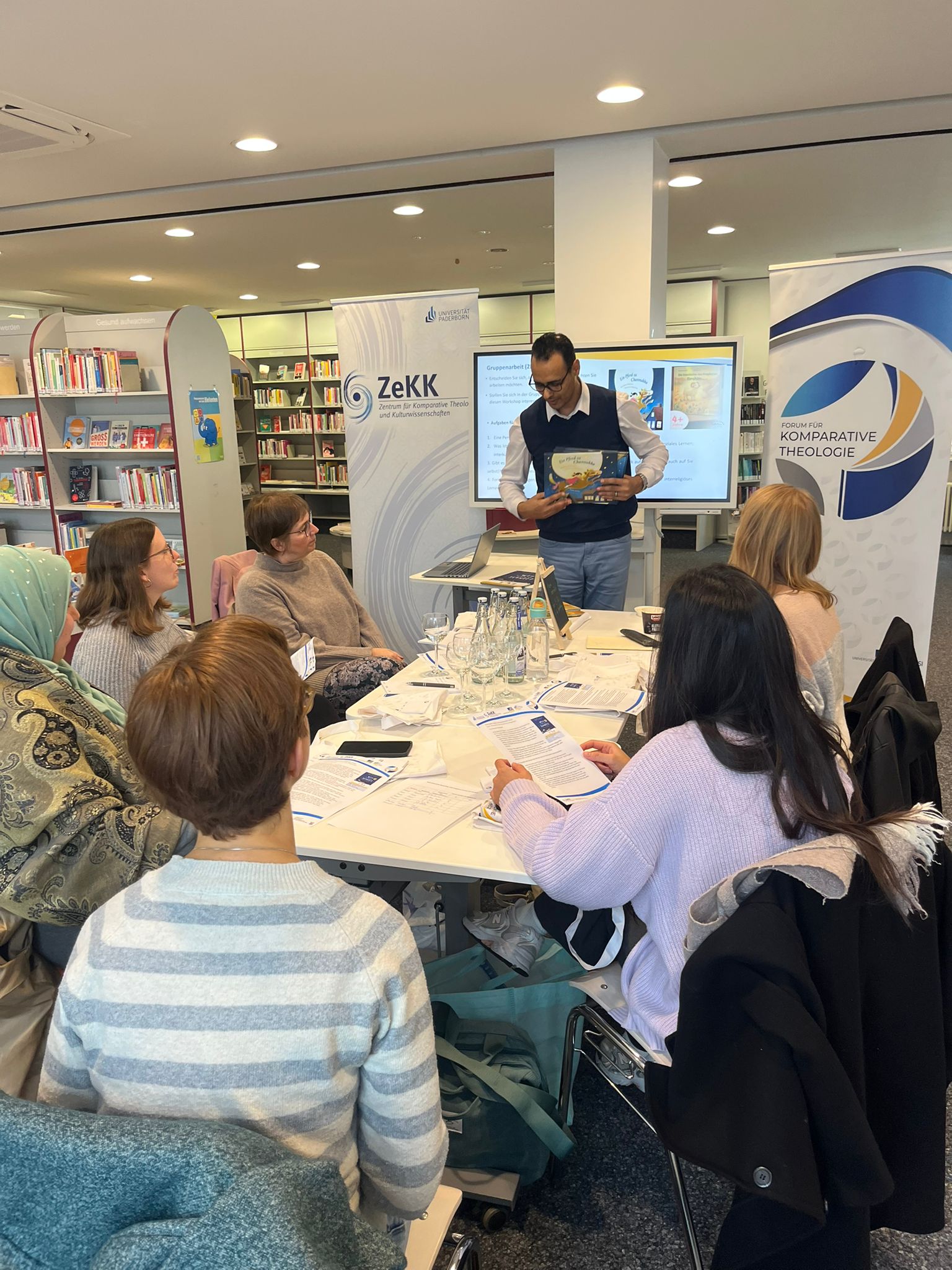 Mehrere Personen sitzen im Kreis um einen Tisch herum Vor ihnen liegen verschiedenen Materialien. Ein Mann steht am Tischende erklärt etwas.