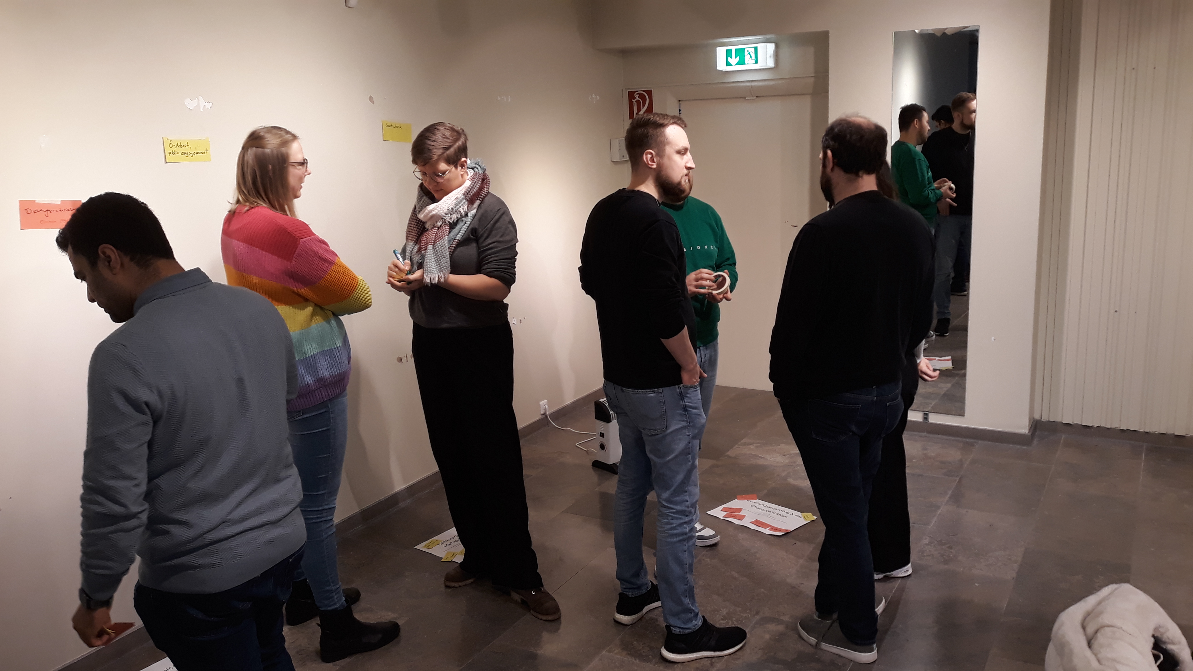 Mitglieder des MAT4HY-Konsortiums bei einem internen Workshop zur Findung neuer Forschungsideen im H2PopUpLab Gelsenkirchen. Die Beteiligten stehen im Raum verteilt und unterhalten sich über Themen, welche durch verschiedene Plakate an den Wänden und am Boden vorgegeben sind. Sie ergänzen ihre Gedanken zu den Themen mittels Post-It-Zettel.