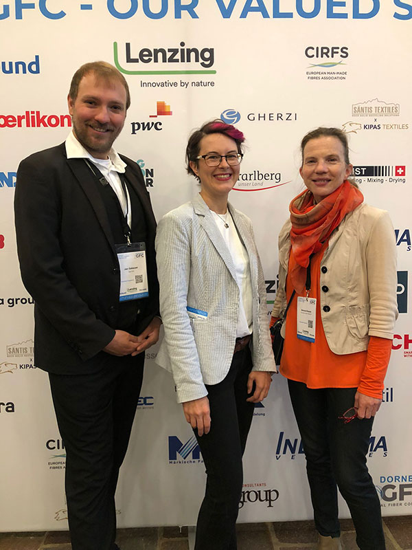 Drei Personen stehen vor einer Wand mit Firmenlogos und lächeln in die Kamera