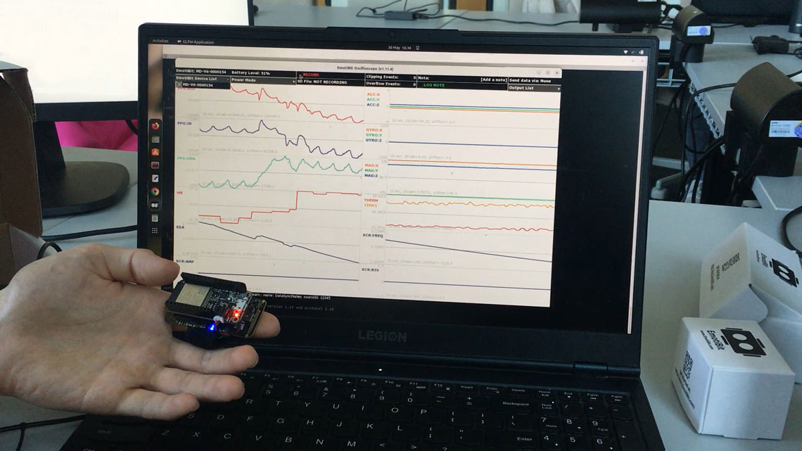 Eine Hand hält ein kleines, mit LEDs bestücktes Elektronikmodul vor einem geöffneten Laptop, auf dessen Bildschirm zeitliche Sensorkurven und Messreihen dargestellt sind.