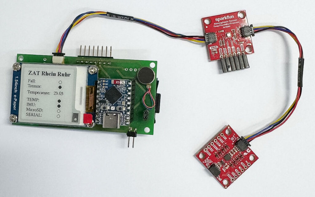 Das Bild zeigt eine elektronische Schaltung auf einer grünen Platine auf der linken Seite. Aufschrift: "ZAT Rhein Ruhr. Fall. Tremor. Temperature. TEMP. IMU. MicroSD. SERIAL." Rechts im Bild sind zwei rote Module zu sehen, die über Kabel mit der grünen Platine verbunden sind.