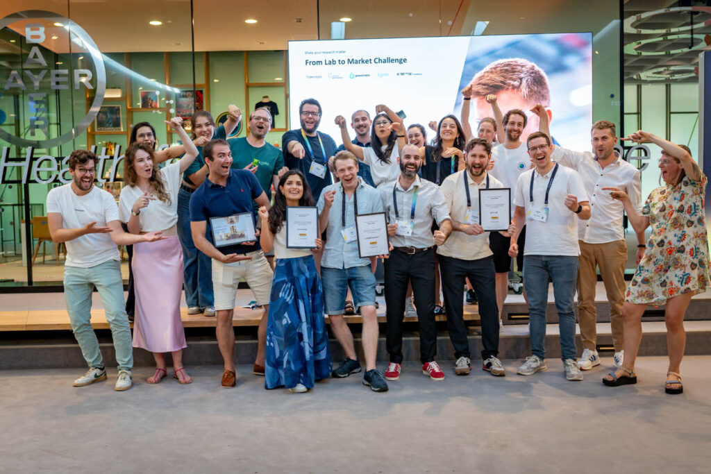 Die teilnehmenden Teams und die Organisatorinnen und Organisatoren der „From Lab to Market Challenge“ 2026 stehen für ein Gruppenfoto zusammen. Die ausgezeichneten Teams halten ihre Zertifikate, während die anderen auf sie zeigen und jubeln. Auf dem Banner im Hintergrund steht „Make your research matter“ und „From Lab to Market Challenge“