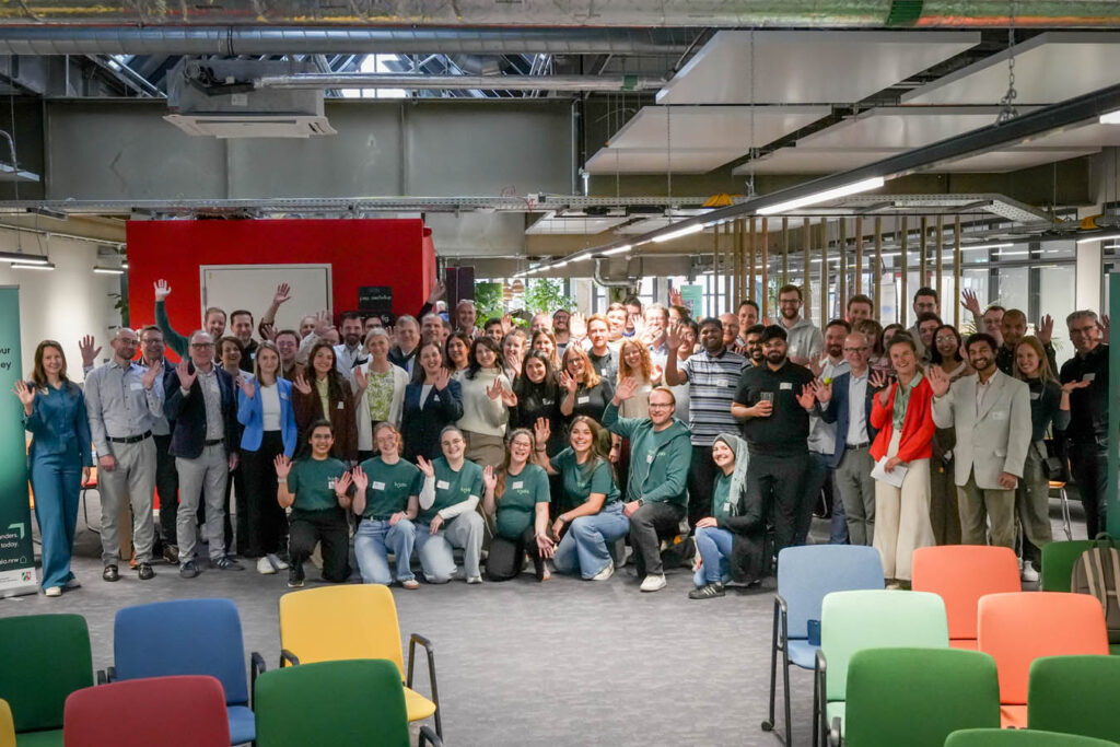 Die Teilnehmenden des Netzwerktreffens kosmos 2026 in Köln stehen für ein Gruppenfoto zusammen und winken in die Kamera. Davor kniet das koala-Team und winkt ebenfalls in die Kamera.