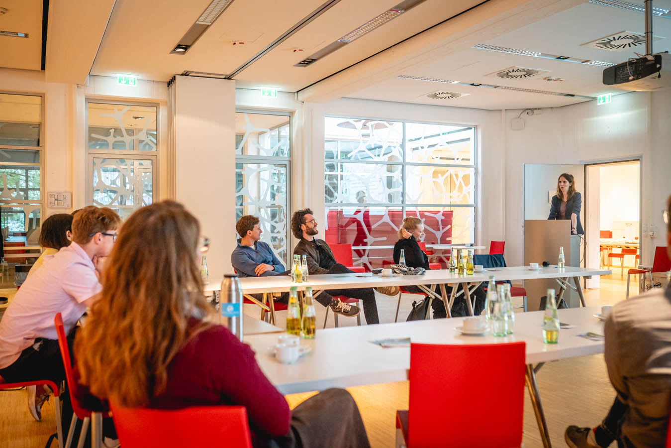 Teilnehmende eines Workshops sitzen an Tischen und hören einer Vortragenden zu.