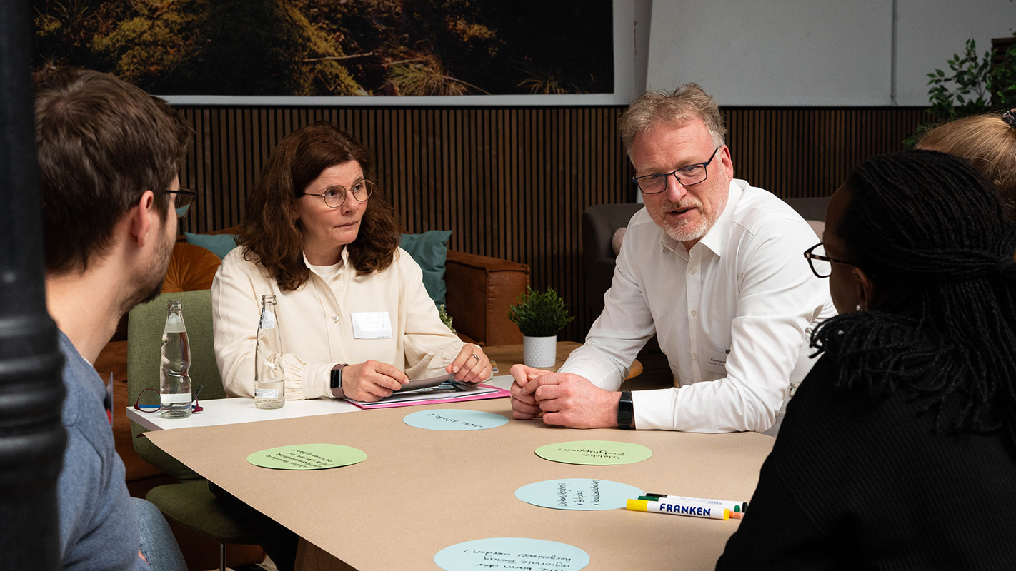 Szene aus einer Arbeitsphase eines Netzwerktreffens von wasser^plus OWL. Mehrere Personen arbeiten gemeinsam an einem Tisch und diskutieren miteinander und beschriften Moderationskarten.